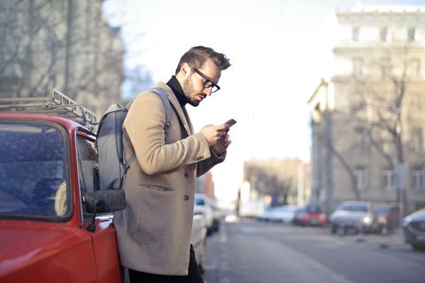 年轻的留着胡子的男性背包客，肩上扛着背包，一边拿着电话，一边靠在红色的汽车上