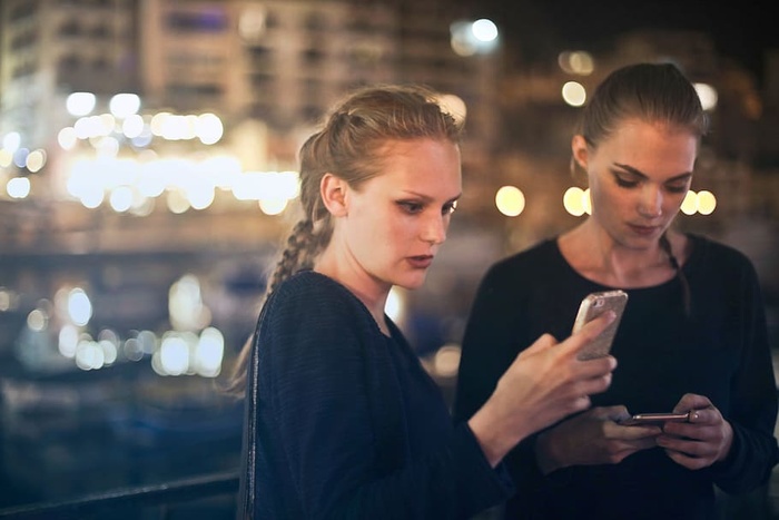 两个穿着黑色连衣裙的金发女郎晚上在户外用手机发短信