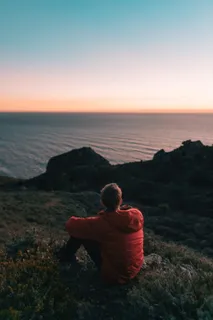 身穿橙色泡泡连帽夹克的男子坐在靠近海洋的地面上