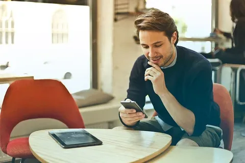 在餐厅里，一个身穿黑色毛衣的年轻人一边用智能手机一边用一只手捂着下巴