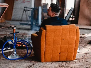 一名男子坐在棕色绒面软垫沙发上，旁边是灰色和蓝色的水烟