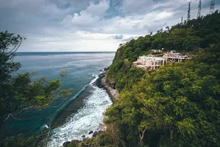 乌云密布的天空下，水体附近绿叶树木之间的白色混凝土建筑