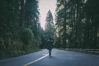 身穿黑色派克大衣的女子在树木环绕的黑色水泥路上行走