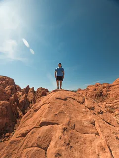 一名身穿蓝色衬衫和黑色短裤的男子站在晴朗天空下的棕色岩层上 一名身穿蓝色衬衫和黑色短裤的男子站在晴朗天空下的棕色岩层上