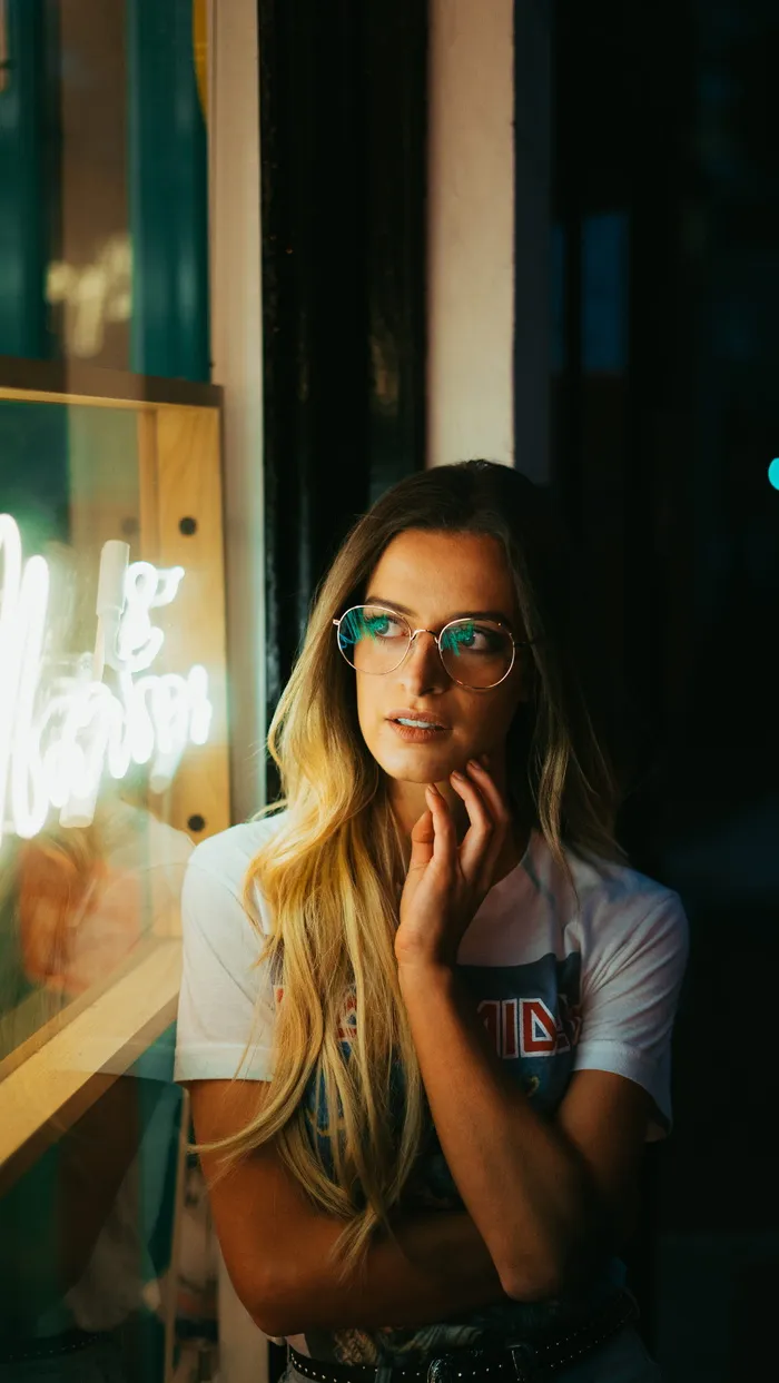 一位戴着眼镜靠在白色墙上在暗室里拍照的妇女