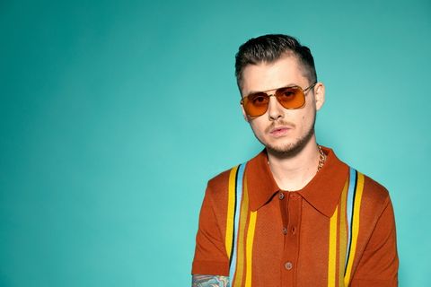 戴墨镜、身穿棕黄色马球衫的男子站在蓝色背景前的肖像照片