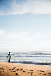 一名年轻的高加索冲浪者在海滩上举着冲浪板,背景是巨浪 一名年轻的高加索冲浪者在海滩上举着冲浪板,背景是巨浪