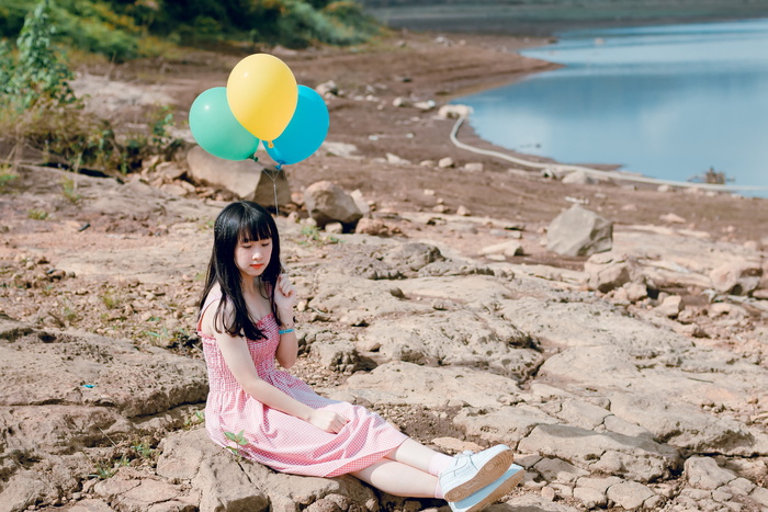 一名身穿粉色厚束带连衣裙的女子坐在岩石地面上，手里拿着一个黄色和蓝色的气球