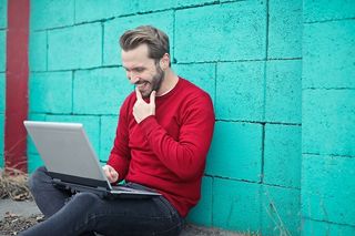 穿着红色毛衣的年轻男子，微笑着用笔记本电脑对着绿松石砖墙