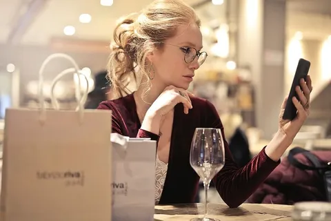 一位身穿缎质连衣裙的金发女郎一边看手机一边喝着葡萄酒 一位身穿缎质连衣裙的金发女郎一边看手机一边喝着葡萄酒
