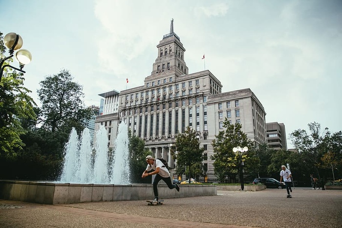 年轻的高加索滑板运动员经过一座建筑物前的喷泉