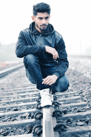 一个穿着黑色皮革拉链连帽衫和黑色牛仔牛仔裤的男人坐在灰色金属火车栏杆上，在沙哑的早晨被岩石包围