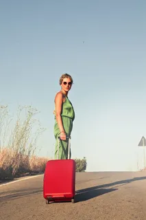 一位身穿橄榄绿连衣裙、戴着太阳镜、拿着红色行李箱的金发女郎摆出姿势 一位身穿橄榄绿连衣裙、戴着太阳镜、拿着红色行李箱的金发女郎摆出姿势