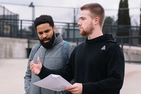 白天,一名男子穿着黑色连帽衫,手里拿着白色打印纸,另一名男子穿着灰色拉链夹克 白天,一名男子穿着黑色连帽衫,手里拿着白色打印纸,另一名男子穿着灰色拉链夹克