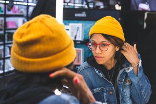 戴着黄色帽子、红色框眼镜、蓝色牛仔夹克、面朝镜子的女人