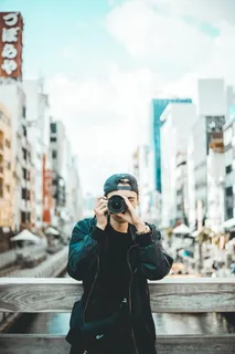 一名男子白天穿着黑色夹克,戴着帽子,手持单反相机 一名男子白天穿着黑色夹克,戴着帽子,手持单反相机