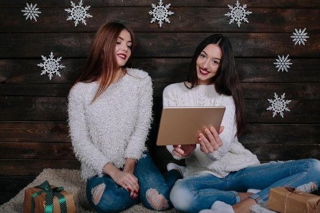 两位美女拿着平板电脑坐在地上，中间夹着圣诞礼物