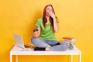 学生在学习或准备考试，感到疲倦和困倦，坐在桌上交叉双腿打哈欠，穿着牛仔裤和绿色t恤隔离黄色背景。