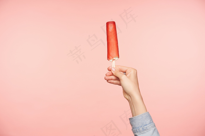 一张年轻女子手拿冰激凌放在涂有红色釉料的棍子上的水平照片，隔离在粉色背景上。食物摄影与人手概念