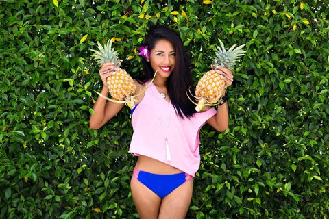 迷人的黑发女孩抱着菠萝，微笑着低头看。一位皮肤黝黑的亚洲女士的户外肖像，蓝色比基尼，紫色花朵，长发，在丛林背景下。