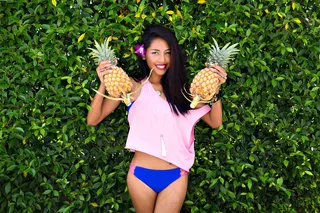 迷人的黑发女孩抱着菠萝，微笑着低头看。一位皮肤黝黑的亚洲女士的户外肖像，蓝色比基尼，紫色花朵，长发，在丛林背景下。