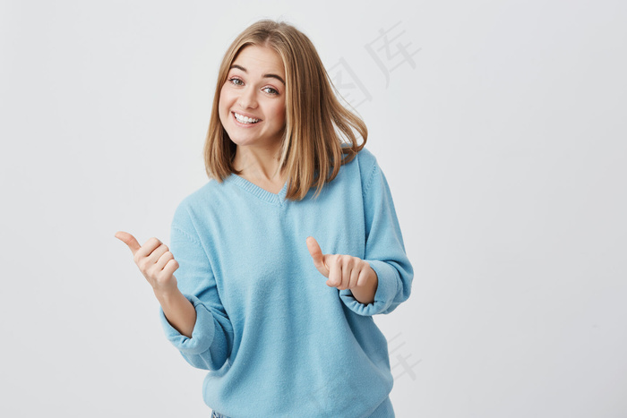 情绪愉快的年轻白人女性，一头金发，穿着蓝色衣服，竖起大拇指，展示了产品有多好。美丽的女孩咧嘴笑着。手势和肢体语言