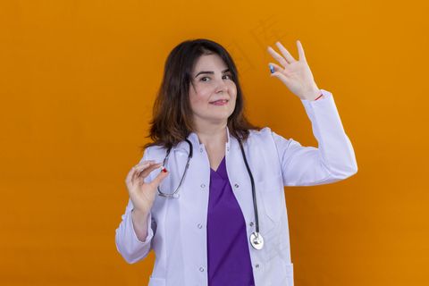 中年女医生，身穿白大褂，手持听诊器，手持药片，站在橙色背景下微笑着看着摄像机
