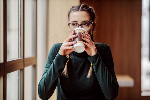 年轻的女大学生坐在靠近窗户的咖啡店里，喝着一次性杯子里的新鲜咖啡。