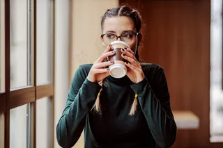 年轻的女大学生坐在靠近窗户的咖啡店里，喝着一次性杯子里的新鲜咖啡。