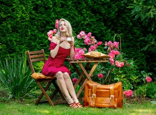 漂亮的金发女孩穿着红裙子，提着手提箱正在花园里喝咖啡