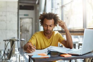 认真专注的年轻非洲大学生穿着黄色t恤，忙着做家庭作业，写在练习本上，一大早就坐在空荡荡的工作空间里，用笔记本电脑