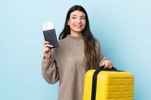 年轻的黑发女孩在隔离的蓝色墙壁上度假时带着手提箱和护照