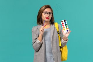 身穿灰色夹克、背着黄色背包、拿着颜料的女学生在蓝色墙上思考的正面图