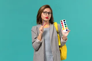 身穿灰色夹克、背着黄色背包、拿着颜料的女学生在蓝色墙上思考的正面图