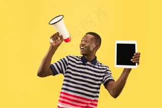 快乐的年轻非洲裔美国人，在黄色的工作室背景上，脸上带着平板电脑。美丽的男性半身肖像。人类情感的概念，面部表情。