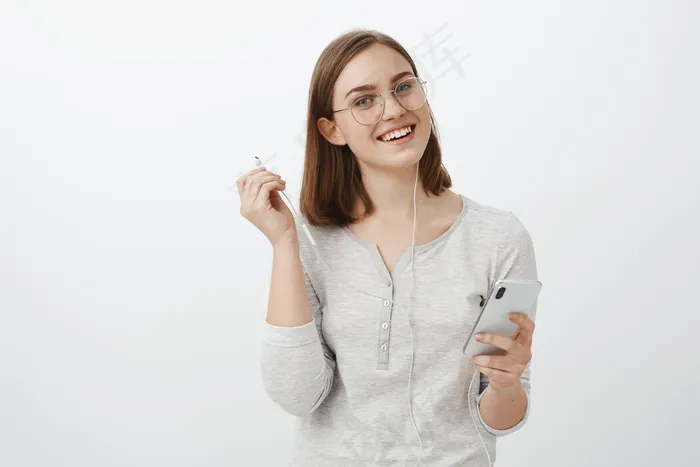 高腰镜头：戴着时髦眼镜的快乐迷人的白种女人，拿着智能手机，摘下耳机，无忧无虑地注视着，听着最喜欢的歌，打断了和朋友的谈话