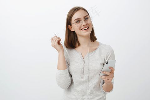 高腰镜头：戴着时髦眼镜的快乐迷人的白种女人，拿着智能手机，摘下耳机，无忧无虑地注视着，听着最喜欢的歌，打断了和朋友的谈话