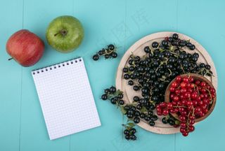 顶视图复制空间红加仑子在一个碗里，黑加仑子在黑板上，笔记本和苹果在浅蓝色的背景上