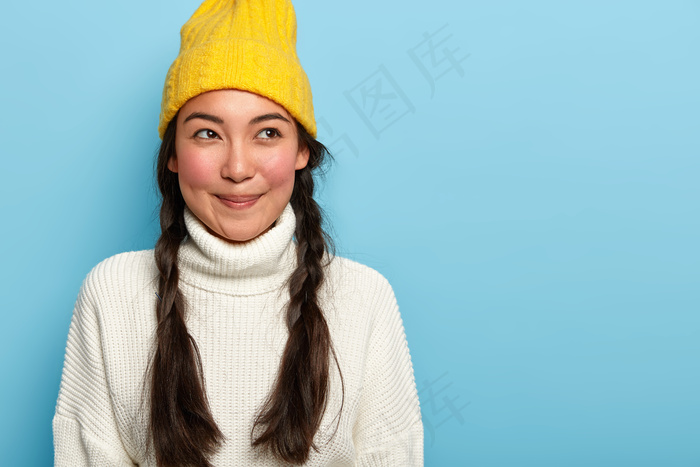 体贴迷人的亚洲年轻女子，笑容宜人，留着两条深色的辫子，身着冬装，蓝色背景下孤立，展现迷人的姿态