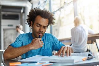 一个卷发、鬃毛的大学生，下巴托着笔，一边看书，一边聚精会神地看书。勤劳的人整天埋头学习