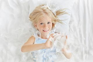 一幅笑容可掬的白人女婴的画像，金发雀斑，躺在床上玩弄着白色的羽毛，美丽稚嫩的脸上洋溢着俏皮欢快的表情