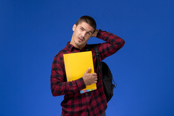 身穿红色格子衬衫、背着黄色文件的男学生在蓝色墙上思考的正面视图