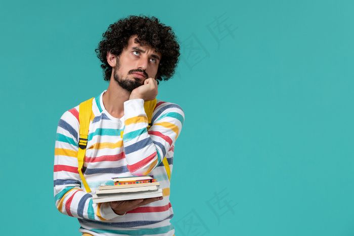 身穿条纹衬衫、背着黄色背包、拿着书的男生在蓝色墙上思考的正面图