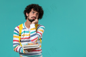 身穿条纹衬衫、背着黄色背包、拿着书的男生在蓝色墙上思考的正面图