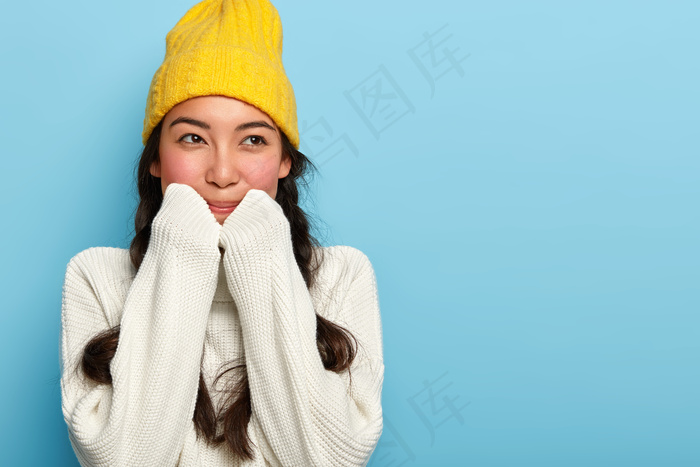 室内拍摄的可爱黑发女子戴着黄色帽子和白色毛衣，有着梦幻般的表情，转眼望去，靠在蓝色的墙壁上