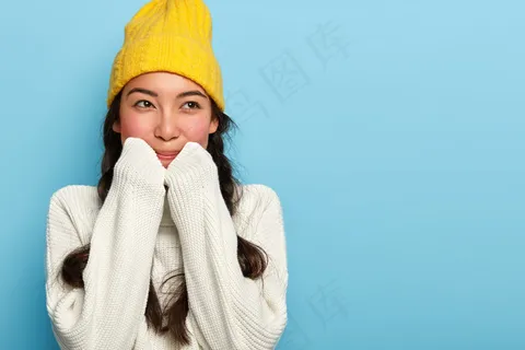 室内拍摄的可爱黑发女子戴着黄色帽子和白色毛衣,有着梦幻般的表情,转眼望去,靠在蓝色的墙壁上 室内拍摄的可爱黑发女子戴着黄色帽子和白色毛衣,有着梦幻般的表情,转眼望去,靠在蓝色的墙壁上