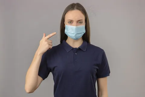 身穿蓝色马球衫,戴着医用防护面罩,手指放在面罩上,神情严肃地站在浅灰色背景上看着摄像机 身穿蓝色马球衫,戴着医用防护面罩,手指放在面罩上,神情严肃地站在浅灰色背景上看着摄像机