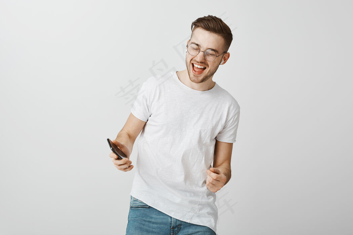 快乐兴奋的家伙微笑着，看着手机，戴着无线耳机跳舞