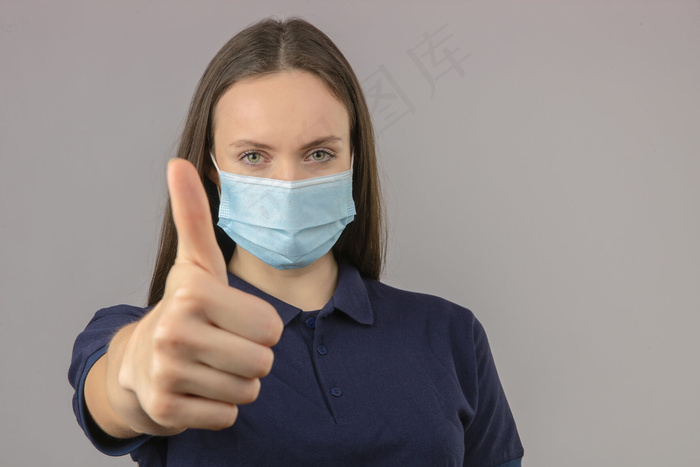 身穿蓝色马球衫的年轻女子，戴防护性医用面罩，站在浅灰色背景上，大拇指朝上，表情积极