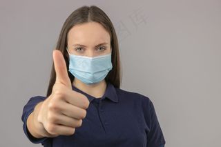 身穿蓝色马球衫的年轻女子，戴防护性医用面罩，站在浅灰色背景上，大拇指朝上，表情积极